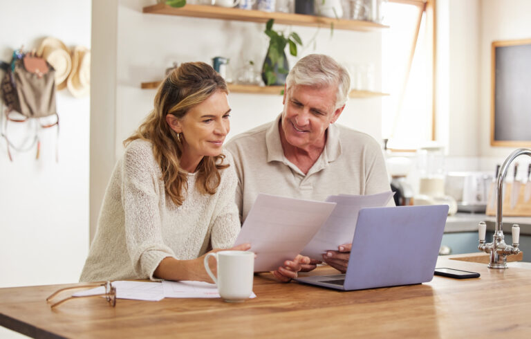 An older couple at their kitchen table, going through paperwork and a laptop together.