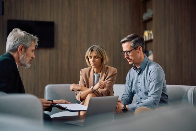 An adviser talking through paperwork with a couple at a table, a laptop and documents in front of them.
